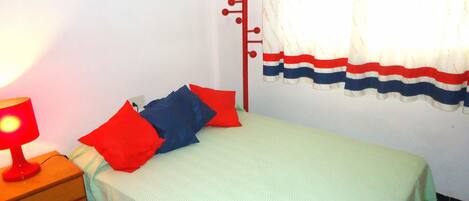 Una cama individual con un edredón verde y dos almohadas, una roja y una azul, en una habitación con paredes blancas y una ventana con cortina a rayas.