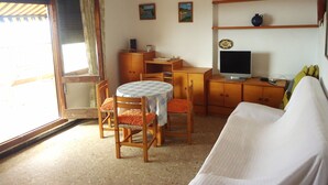 Ein Zimmer mit einem Holztisch, einem Fernseher, einer Couch und einem Esstisch mit Stühlen. An der Wand hängen zwei Bilder und es gibt ein Fenster mit Vorhängen.