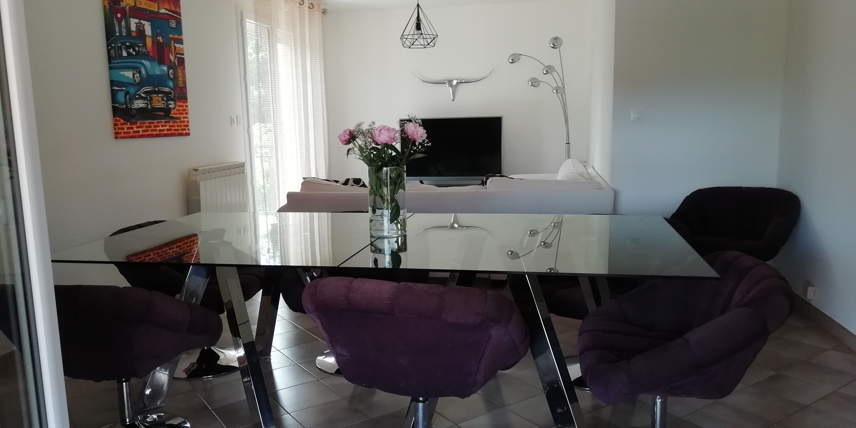 Un espace repas moderne avec une table en verre, des chaises violettes, un vase avec des fleurs, un téléviseur et un élément décoratif mural.