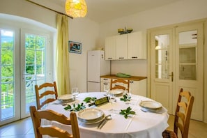 Uma área de refeições com uma mesa redonda posta para quatro pessoas, rodeada por cadeiras de madeira. Há um armário branco com um tecido verde em cima, uma geladeira branca e uma porta com painéis de vidro.