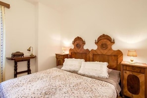 Quarto com cabeceira de madeira, duas mesas de cabeceira, colcha estampada e mesa de canto com abajur.