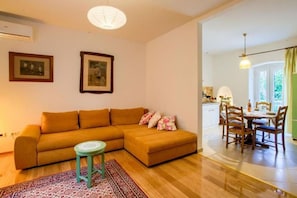 Sala de estar com sofá, mesa redonda pequena e tapete estampado. Quadros emoldurados adornam a parede e há uma área de jantar com cadeiras e uma mesa.