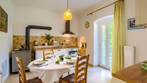 A dining area with a round table set for four, wooden chairs, a hanging light fixture, a kitchen in the background, and a view of the outdoors through a glass door. - Attractive holiday home in Kraljevica with garden (Kraljevica)