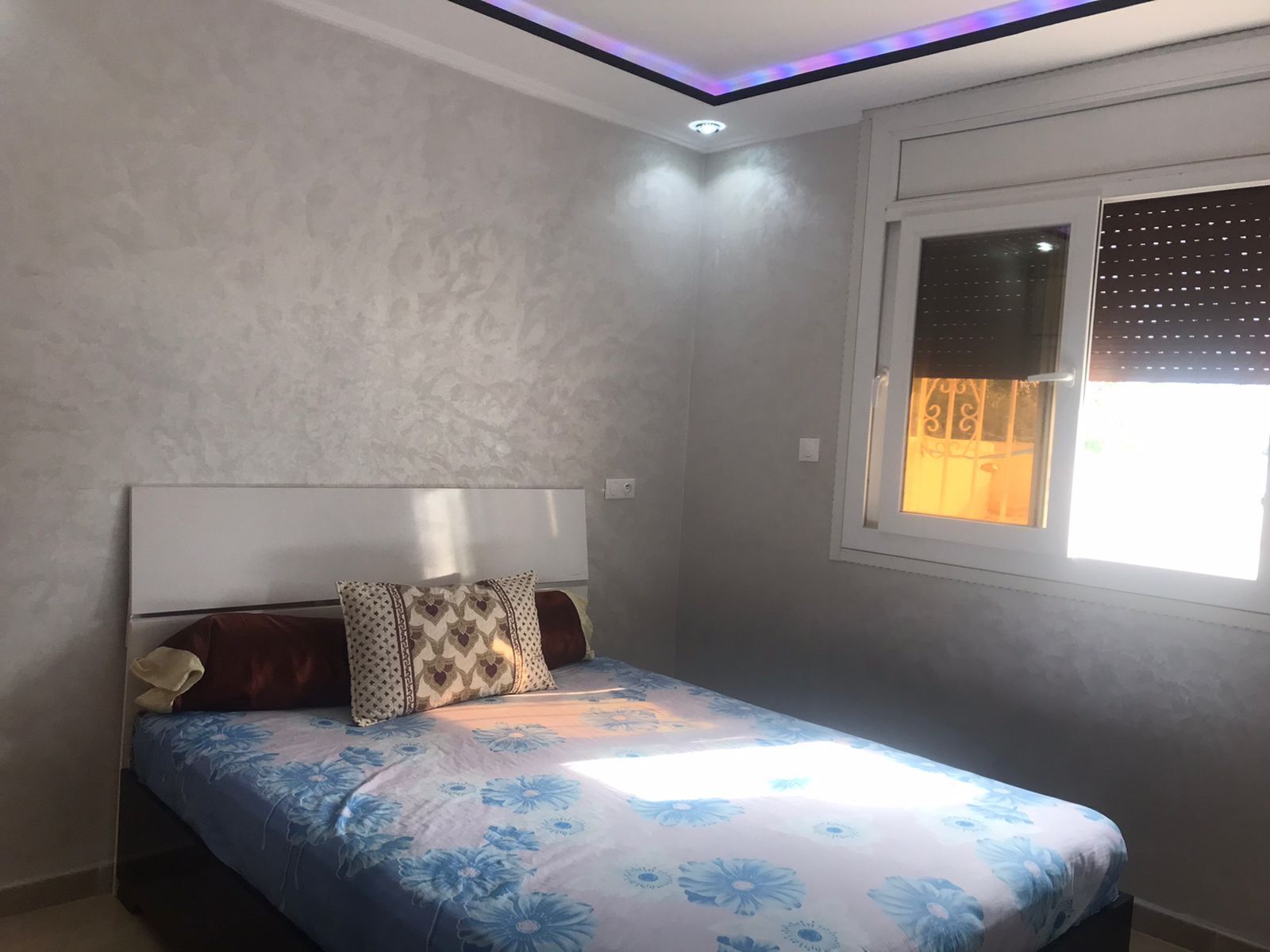 Ein Schlafzimmer mit einem Bett, Kissen mit Muster, einem Fenster mit Jalousien und einer Deckenleuchte.