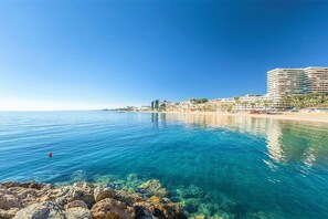 Vista costera con aguas azul turquesa, un litoral rocoso y un hotel frente a la playa con varios pisos y balcones.