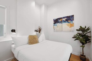 Une chambre à coucher avec un lit, des murs blancs, une fenêtre, une plante et un tableau.