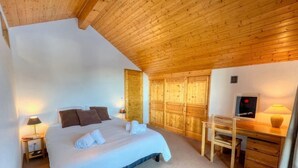 A bedroom with a wooden ceiling, a bed with white bedding, a desk with a chair, and a poster on the wall.
