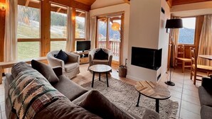 A cozy living room with a fireplace, comfortable seating, and large windows offering a view of the outdoors.