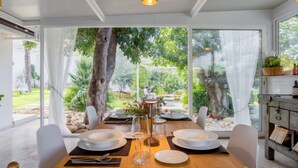 A dining area with a wooden table set for four, large windows offering a view of a garden with trees and a patio.