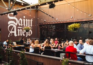 Un espace extérieur aménagé pour les repas, avec des tables et des chaises, une grande enseigne indiquant « Saint Anejos » et des plantes suspendues.