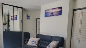 Un salon avec une table ronde, des fauteuils gris, un canapé noir orné de coussins à motifs et un tableau accroché au mur.