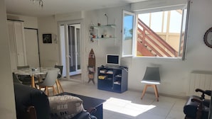 Un salon comprenant un canapé, une table basse, un téléviseur et une bibliothèque. Une porte donne sur un balcon d’où l’on peut admirer un immeuble.
