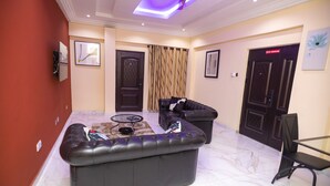 A living room with two black sofas, a glass coffee table, a ceiling fan, and a 'No Smoking' sign on a door. - 2 Bed Apartment - Parking, Wifi, Aircon (Accra)