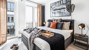 Une chambre moderne avec un grand lit, une table de chevet avec une lampe, un plateau en bois avec des verres et une affiche représentant un paysage urbain accrochée au mur.