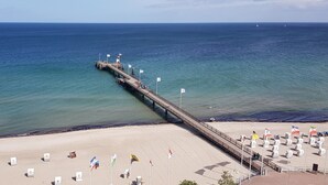Une jetée qui s’avance dans la mer, avec une plage aménagée de chaises longues et de drapeaux.