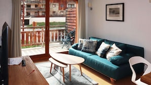 A living room with a teal sofa, a wooden coffee table, and a TV on a wooden stand. There is a balcony with a view of a building and a fence. - Ski-in/Ski-out Apartment with Pool (6 pers) - Les Orres 1800 (Les Orres)