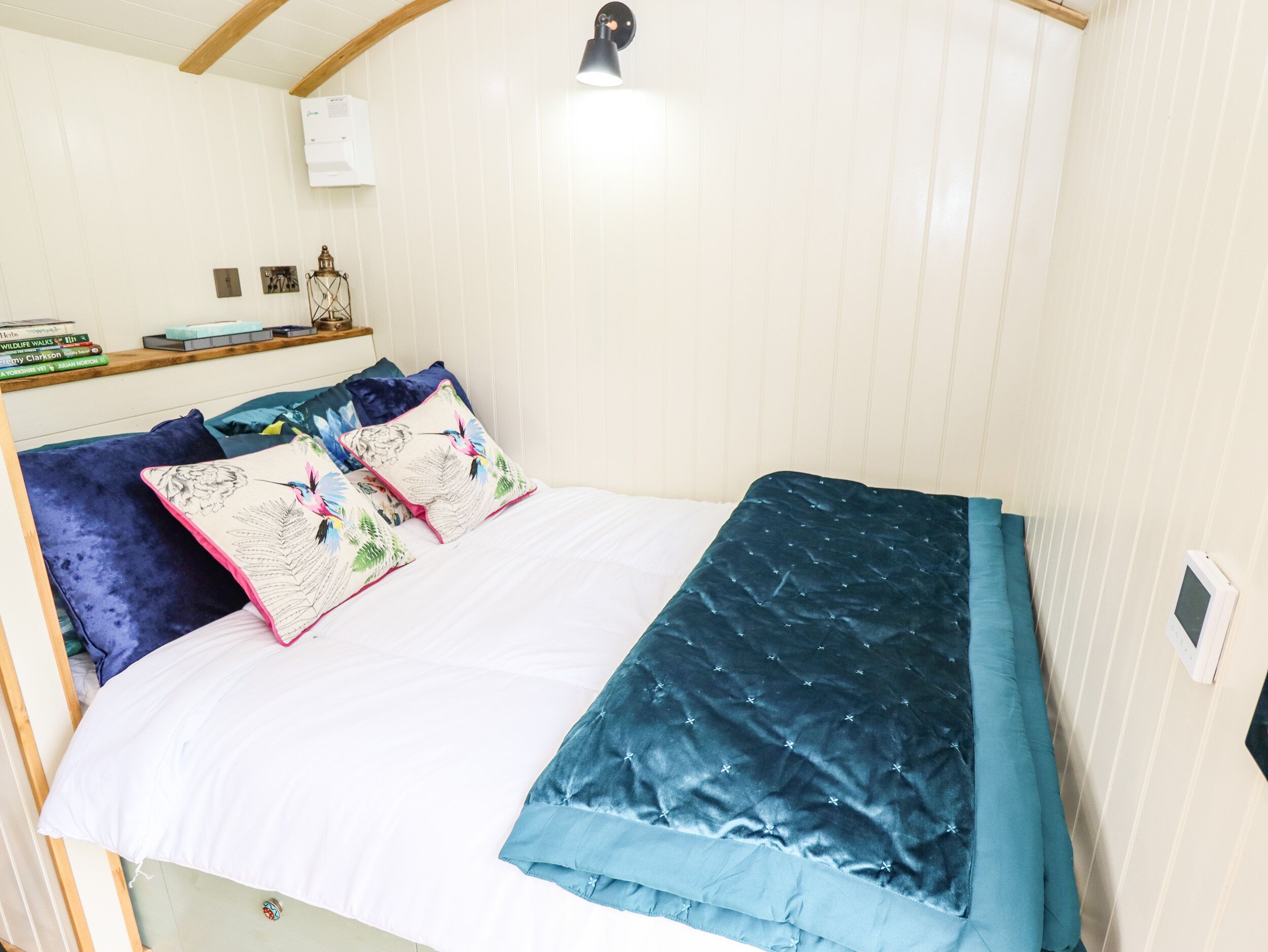 Una cama bien hecha con edredón blanco y azul, dos almohadas decorativas con diseños florales, una colcha azul oscuro y una pequeña mesita de noche con una lámpara y libros.