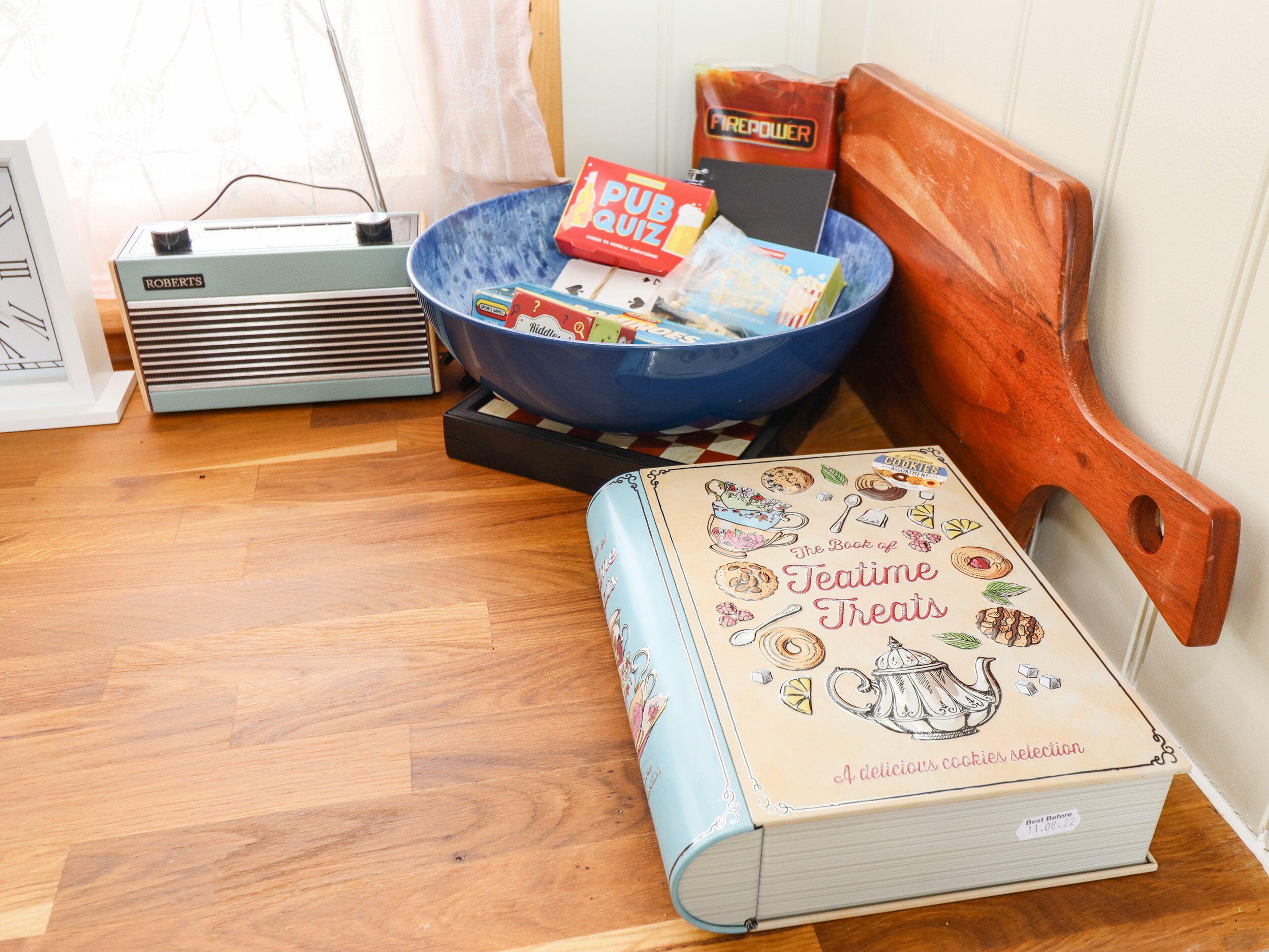 Una stanza con pavimento in legno, una ciotola blu piena di oggetti assortiti, una radio vintage, un libro intitolato 'The Book of Teatime Treats' e una sedia di legno.