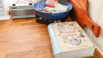 Una stanza con pavimento in legno, una ciotola blu piena di oggetti assortiti, una radio vintage, un libro intitolato 'The Book of Teatime Treats' e una sedia di legno.