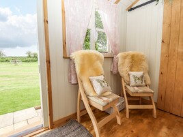 Dos sillas de madera con cojines de piel y almohadas de mariposa, ubicadas cerca de una ventana con cortinas traslúcidas, con vistas a un espacio exterior cubierto de césped.