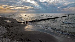 Una playa con un muelle de madera que se adentra en el mar, olas suaves rompiendo en la orilla y un cielo nublado.