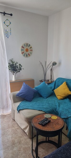 A living room with a beige sofa, a blue and yellow blanket, a small round table with a remote control, a decorative clock, and a plant. - "EL COTILLO TRANQUILIDAD"/Stylish/Minutes from swim or surf/Well equipped (El Cotillo)