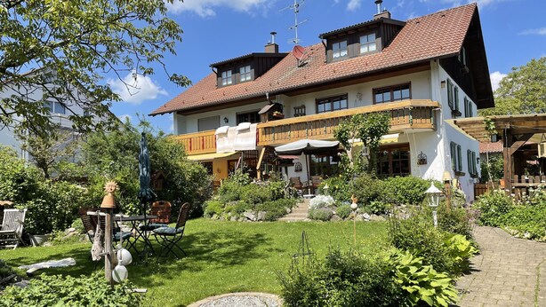 Ein großes, zweistöckiges Gebäude mit einem Vorgarten, der einen gepflasterten Innenhof, Sitzgelegenheiten im Freien und verschiedene Pflanzen umfasst.