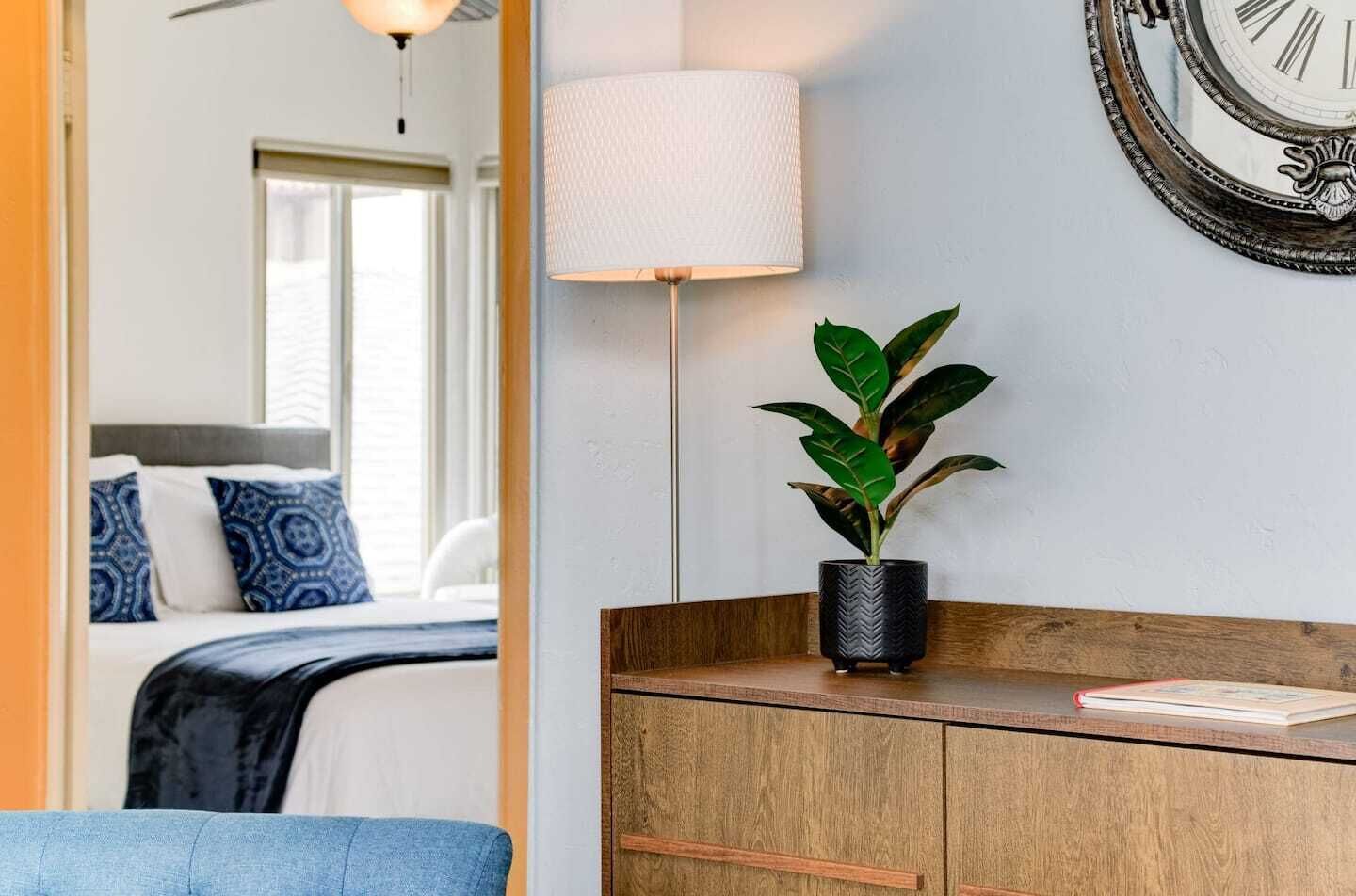 Une chambre d’hôtel avec un lit, une lampe de chevet, une plante en pot, une armoire en bois et une horloge murale.
