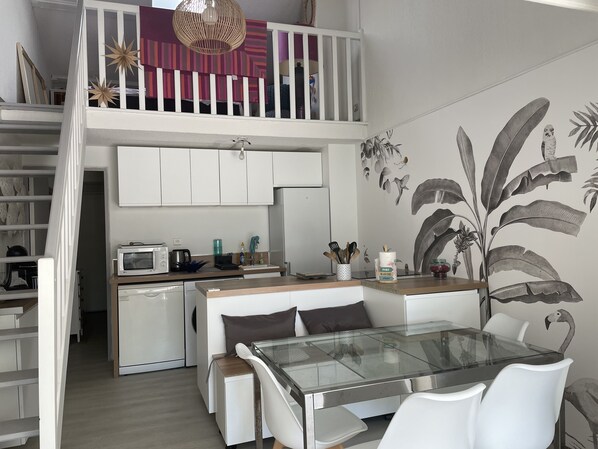 A modern kitchen with a glass dining table, white cabinets, and a loft bedroom above. - Renovated, air-conditioned house, 5 minutes from the beach (Saint-Cyr-sur-Mer)