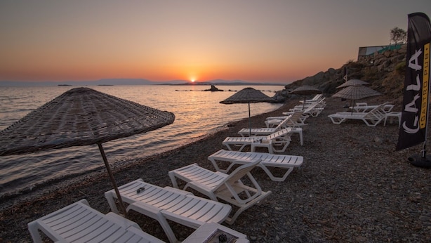 Une plage avec des chaises longues blanches et des parasols en chaume, un coucher de soleil en arrière-plan, et un drapeau sur lequel on peut lire « Castro Pub ».