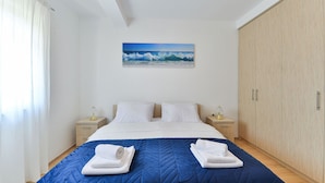 Una cama bien hecha con un edredón azul, dos almohadas blancas y dos toallas dobladas sobre la cama. Hay un cuadro de una ola en la pared sobre la cama.
