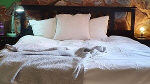 Una cama bien tendida con sábanas blancas y un edredón mullido, un muro de piedra al fondo y una mesita de noche con una lámpara y una pequeña botella.