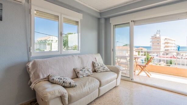 A room with a beige sofa, two windows, and a balcony with a view of buildings. - "El Cid" Holiday Apartment with Sea View, Pool, Garden & Terrace (Denia)