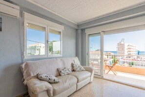 A room with a beige sofa, two windows, and a balcony with a view of buildings.