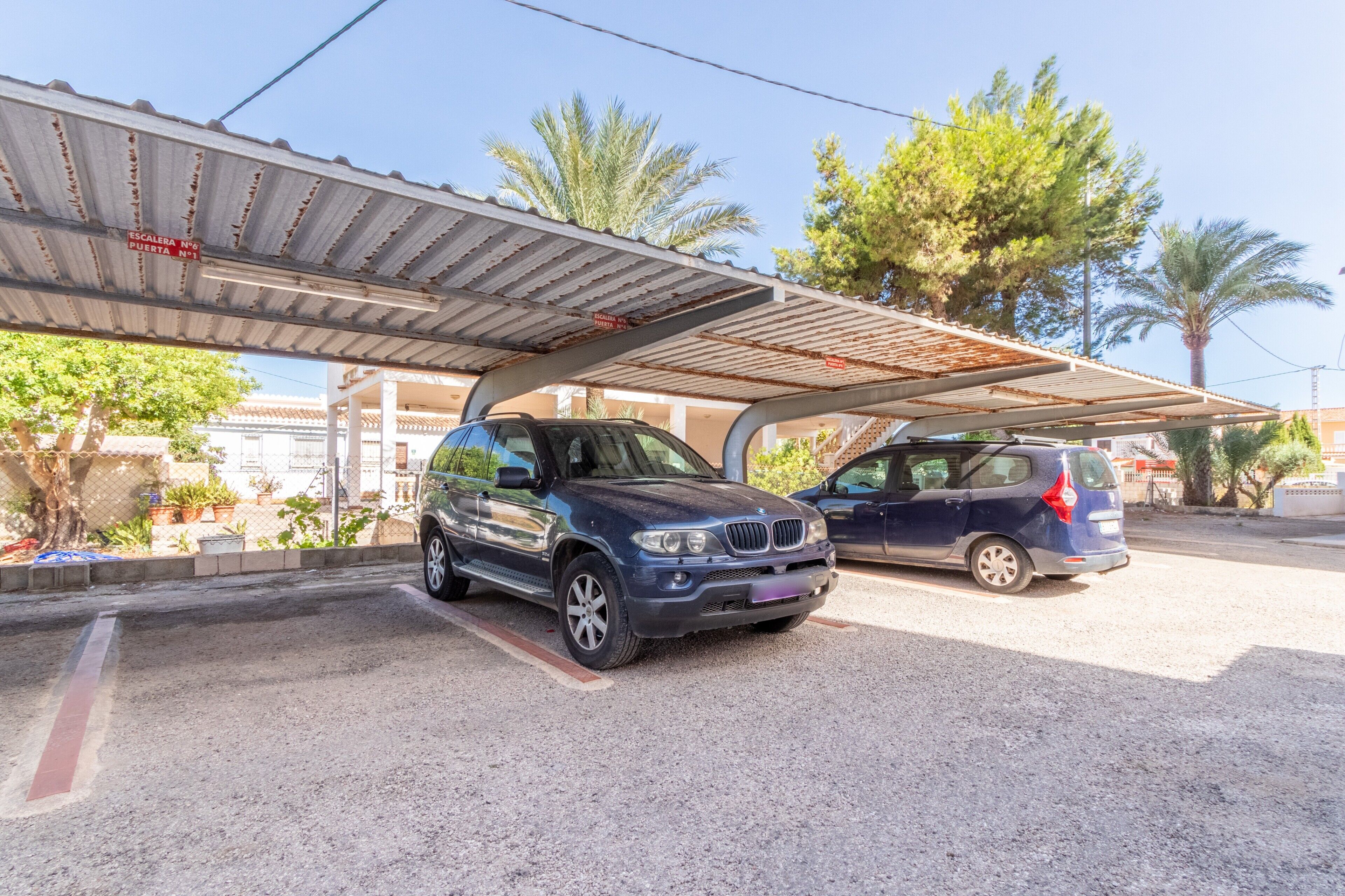 A parking area with two cars, a covered structure, and palm trees in the background.