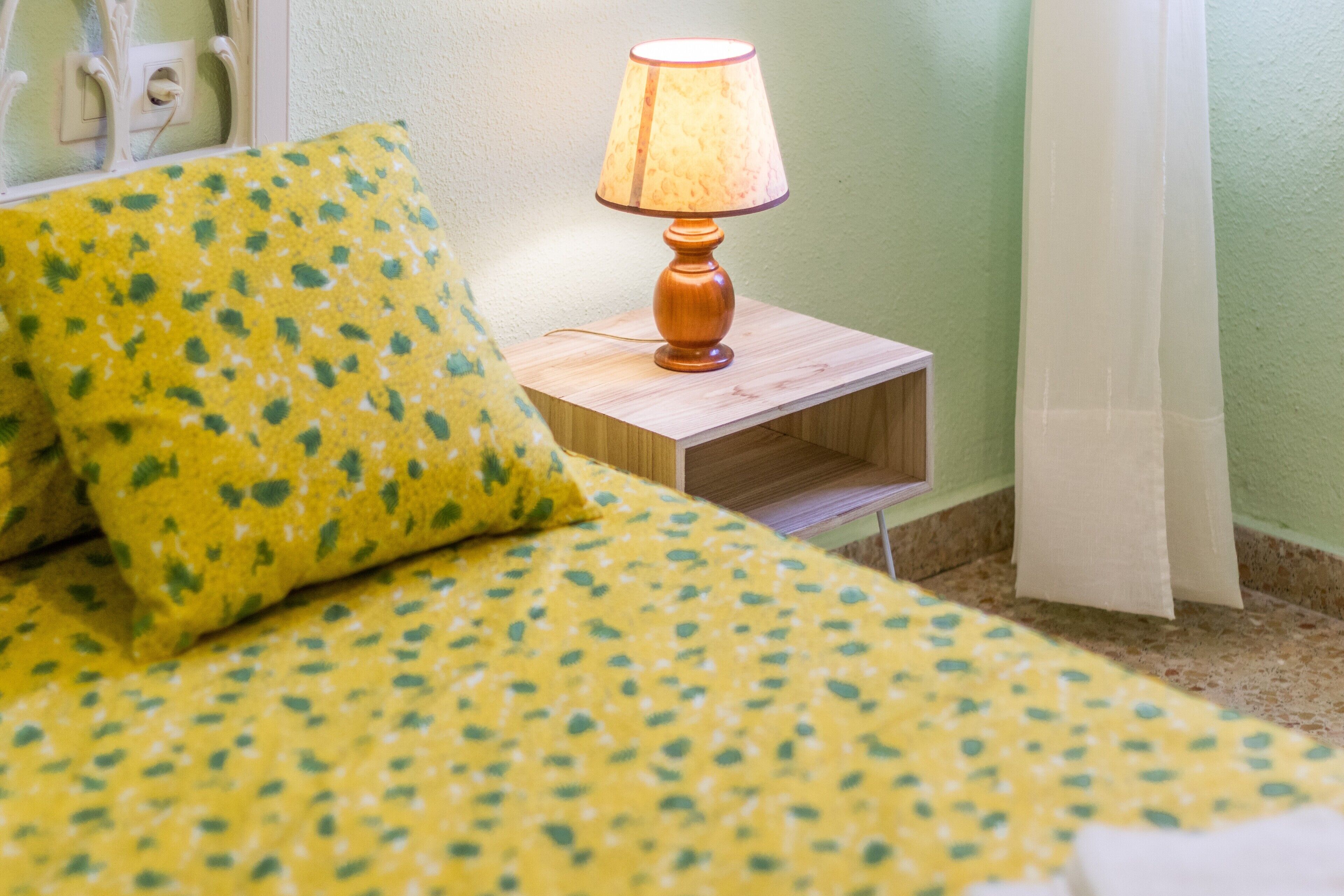 Un lit recouvert d’une couvre-lit à motifs floraux jaunes, agrémenté d’un coussin assorti, une table de chevet en bois avec une lampe, et un mur vert.