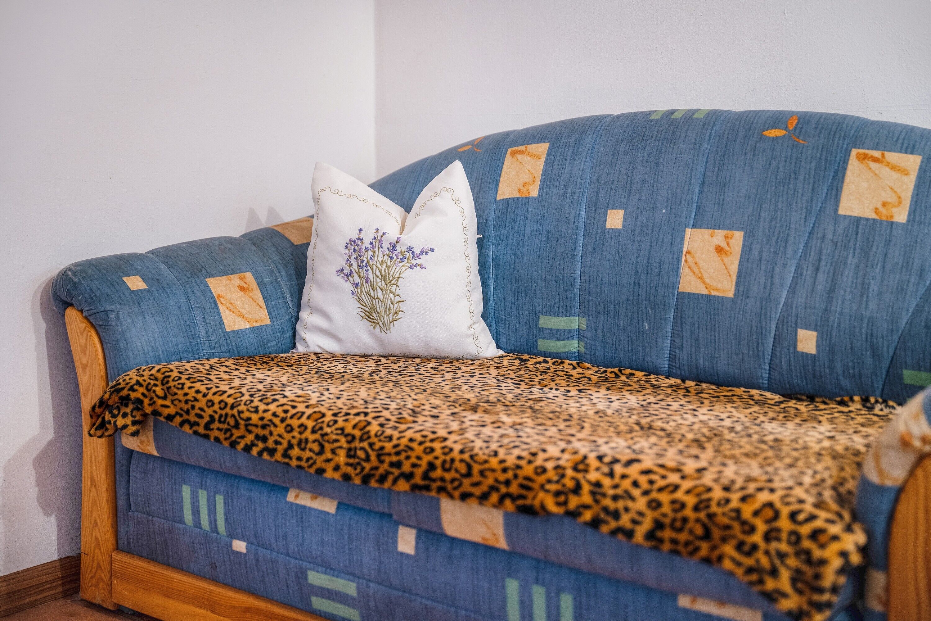 Un sofá azul con un plaid de estampado de leopardo, una almohada blanca con diseño floral y un marco de madera.