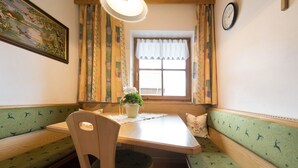 A room with a wooden table, a bench with a green pattern, a potted plant, and a window with curtains.