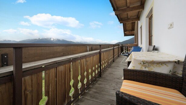A balcony with a wooden railing, a wicker chair with a striped cushion, and a table with a floral tablecloth.