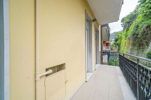 A narrow balcony with a yellow wall, a black metal railing, and a view of a hillside with greenery.