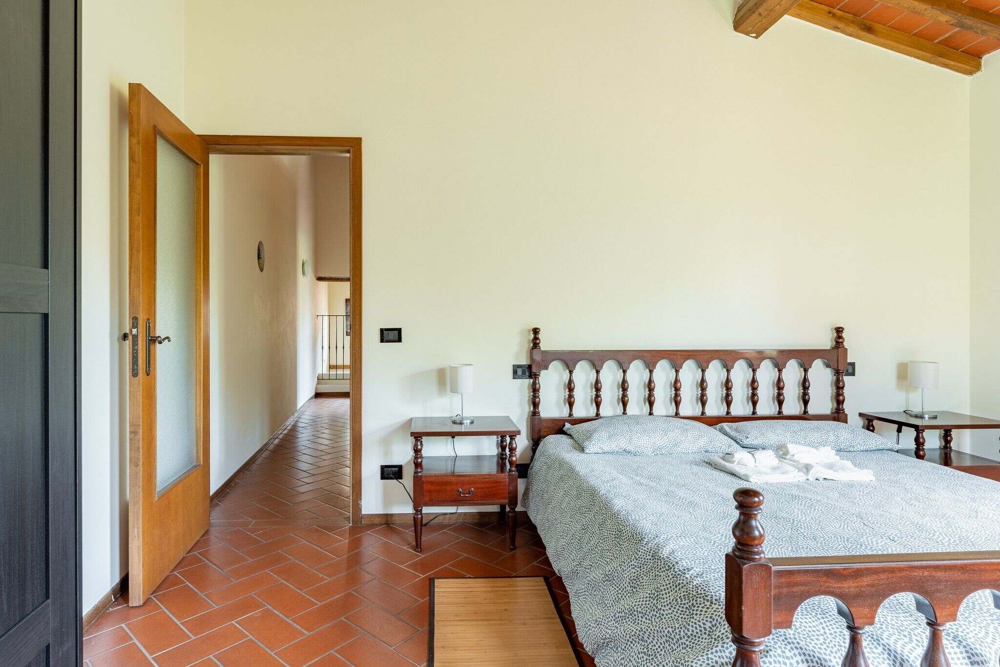 A bedroom with a wooden bed, two bedside tables, a nightstand, a tiled floor, and a door leading to another room.