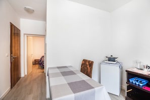 A compact room with a bed, a small refrigerator, a wooden chair, and a desk with a kettle and a coffee maker.