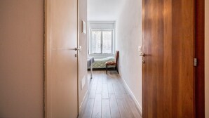 Um corredor estreito com uma porta de madeira à direita, uma janela com vista e uma cama com cobertor verde ao fundo.