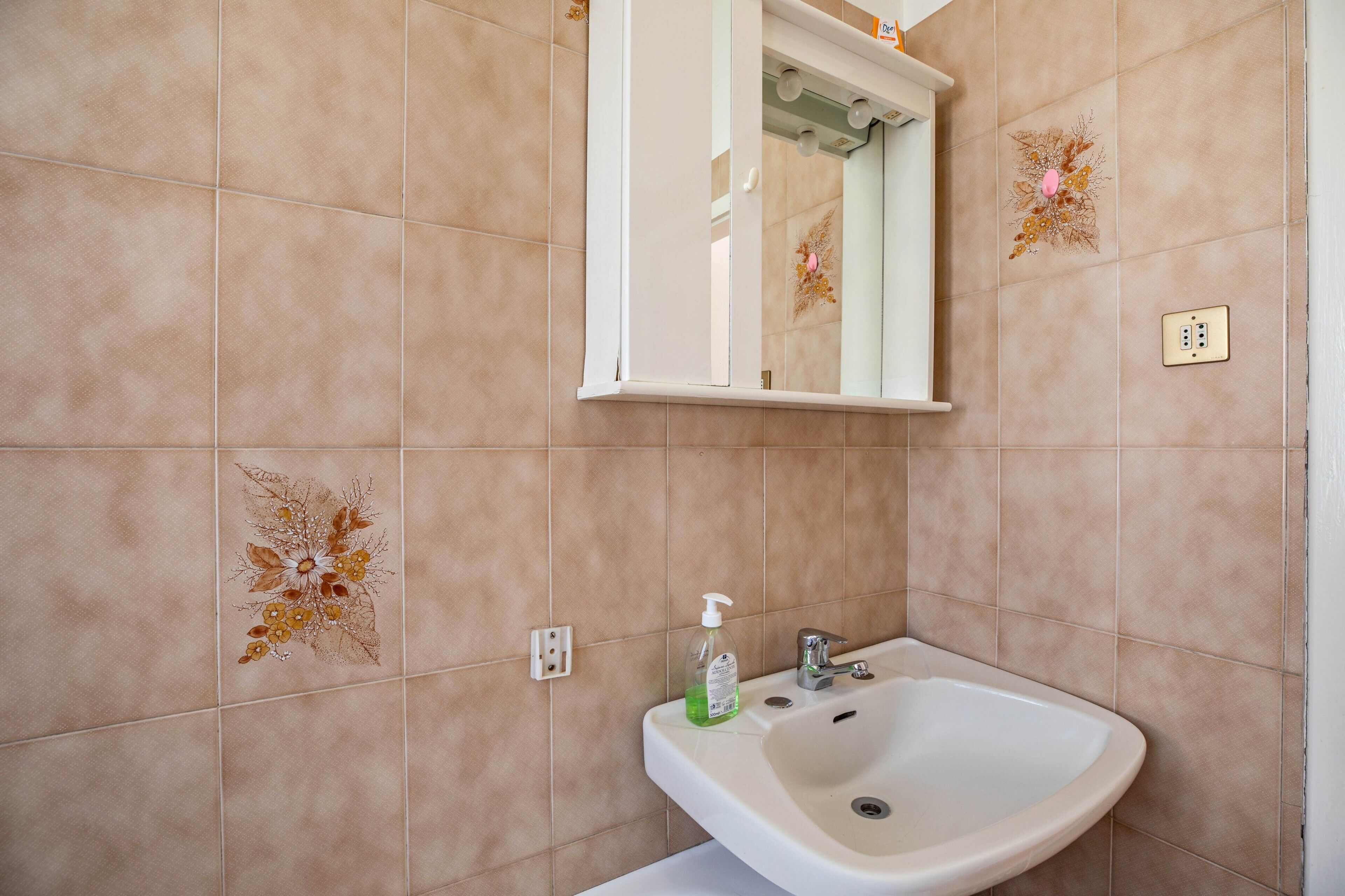 Ein Badezimmer mit beigen Fliesen, einem weißen Waschbecken, einem Spiegel und dekorativen Blumenfliesen.