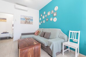 A living room with a sofa, a wooden coffee table, a white chair, a wall with decorative plates, and a painting. - Vacation Home Casa Eri close to the Beach with Pool, Terrace & Wi-Fi (Corralejo)