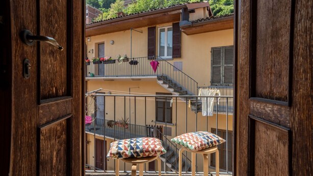 An open wooden door revealing a balcony with two stools and cushions, a railing, and a view of a building with balconies and greenery. - GuestHost - Cozy, completely renovated 47sqm apartment located on the 3rd floor of a building WITHOUT elevator. The apartment is accessed via an external balcony overlooking the quiet internal courtyard.The accommodation is located in an excellent locati (Como)