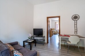 A modern living room with a sofa, a TV, a small table with a plant, and a dining area with a desk and chairs.