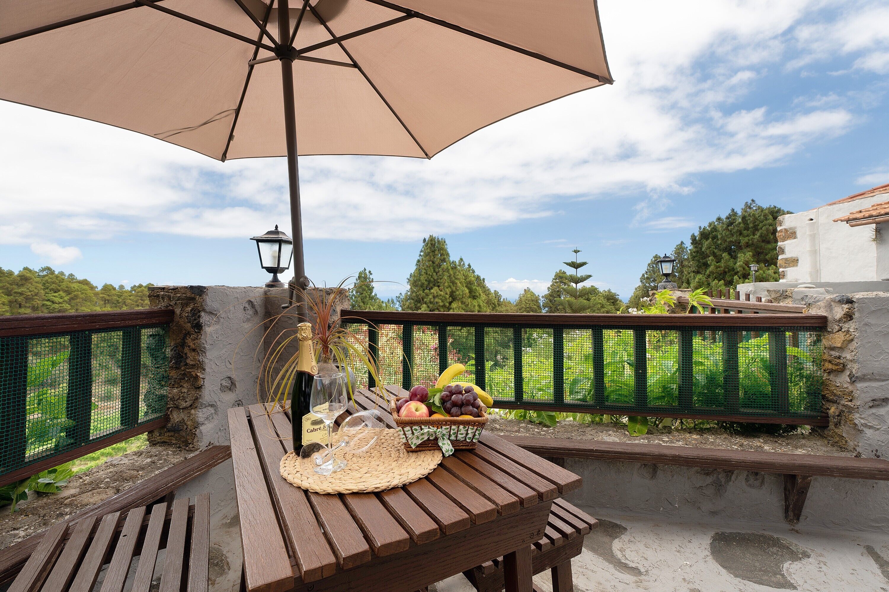 Een houten tafel met een geweven tafelmat, een rieten mand vol fruit, een fles champagne en een glas wijn onder een grote beige parasol.