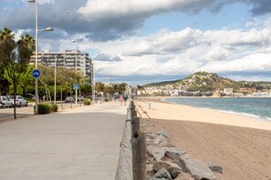 A coastal walkway with a beach, a rocky barrier, and a hill with buildings in the background.