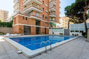 Une piscine avec un bâtiment en briques en arrière-plan, une entrée fermée par un portail et quelques chaises longues.